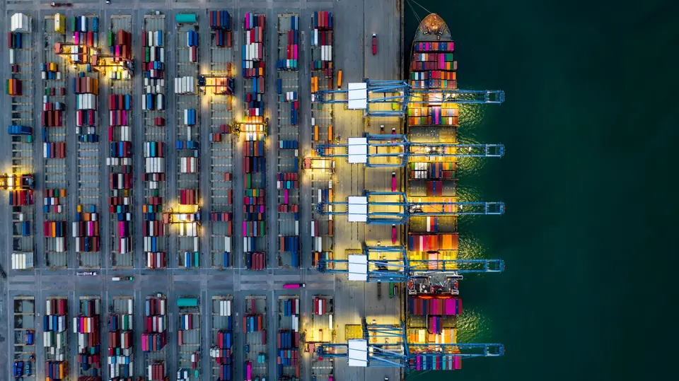 Containerterminal bei Nacht von oben: beleuchtete Stellflächen voller Container, am Kai ein Containerschiff